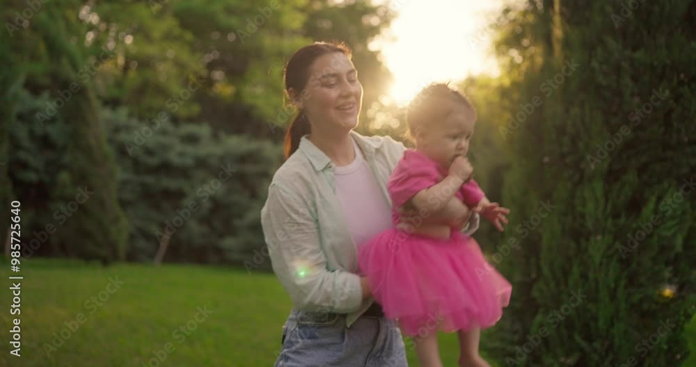 Happy brunette girl in shirt swinging her little daughter in pink dress in her arms during happy walk in park on lawn Happy brunette girl in shirt swinging her little daughter in pink dress in her arms during happy walk in park on lawn
