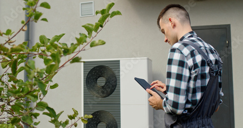 A technician uses a tablet to adjust settings on an external heat pump unit outside a home. photo