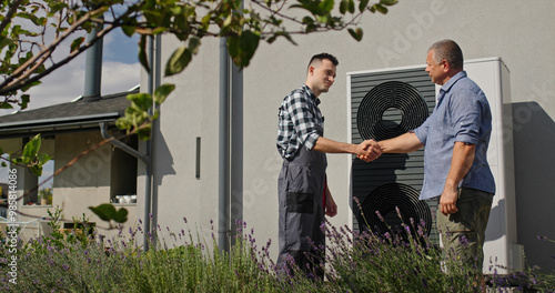 A technician and a homeowner shake hands beside an external heat pump, concluding a service visit.