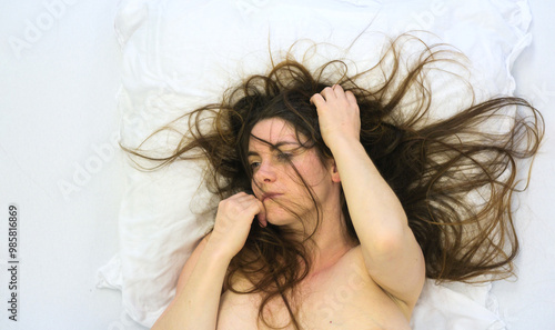 Billede på lærred sexy nude woman with disheveled hair lies on a white pillow, her expression distant and reflective