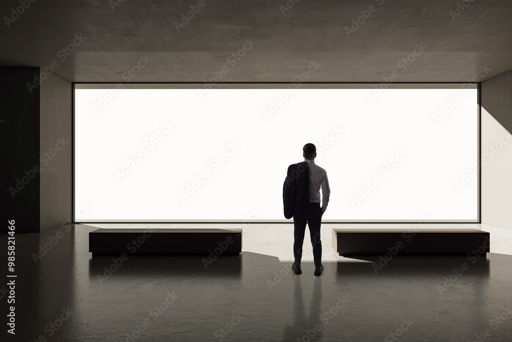 Businessman standing in a modern empty room with large window, looking outside in contemplation. Businessman standing in a modern empty room with large window, looking outside in contemplation.