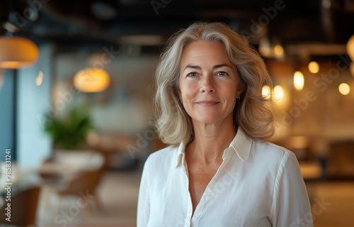 A confident woman with wavy gray hair smiles warmly in a well-lit, modern interior setting.