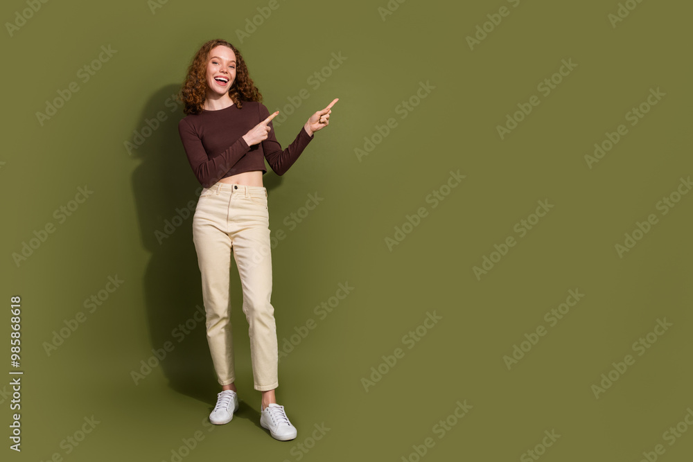 Full length photo of charming positive girl wear brown shirt pointing two fingers empty space isolated green color background Full length photo of charming positive girl wear brown shirt pointing two fingers empty space isolated green color background