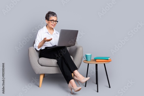 Full body photo of attractive pensioner woman sit armchair hold netbook video call dressed formalwear isolated on gray color background