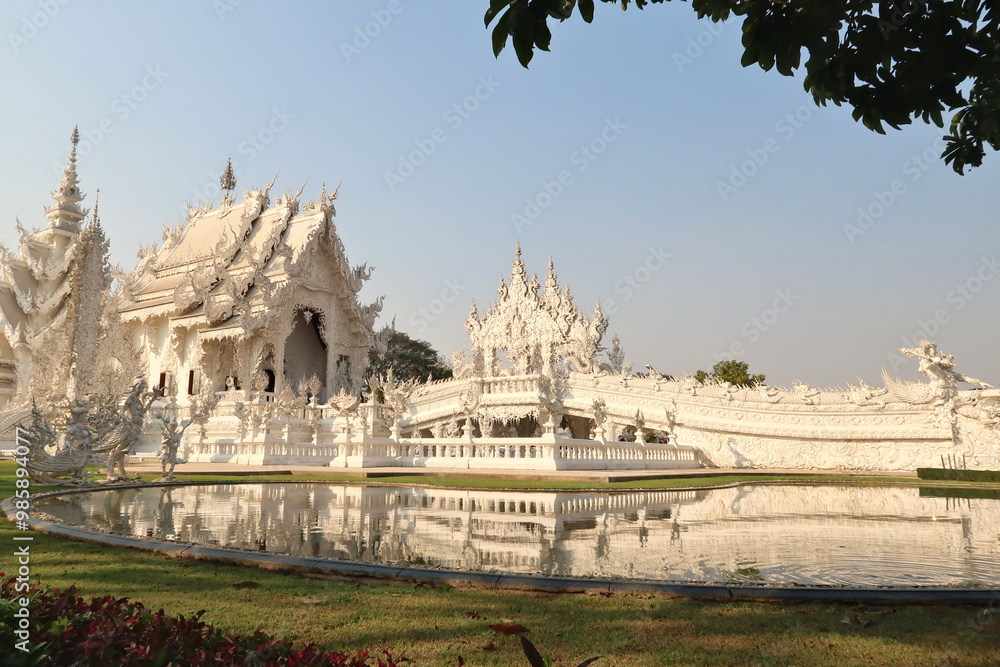 Fototapeta premium View onto the White Temple, Wat Rong Khun, the temple and the bridge leading to it are reflecting in the pond, lake in front of it, Chiang Rai, Thailand