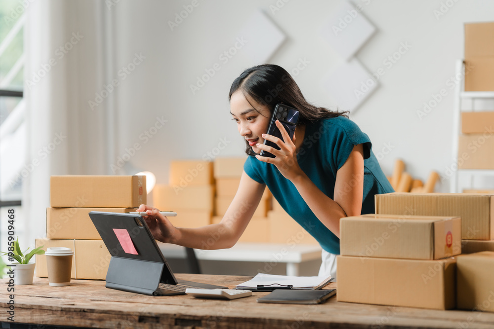 E-commerce Hustle: Young Asian entrepreneur juggling calls and orders amidst a flurry of packages in her bustling home office. E-commerce Hustle: Young Asian entrepreneur juggling calls and orders amidst a flurry of packages in her bustling home office.