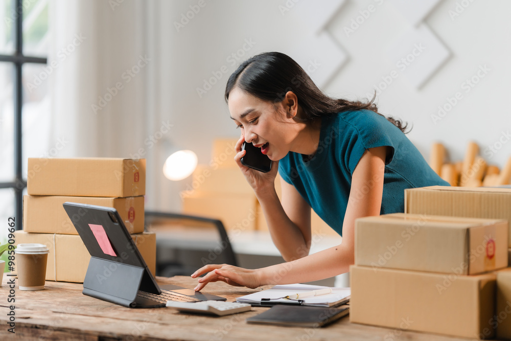 E-commerce Hustle: Young Asian entrepreneur managing orders and logistics on phone and tablet, surrounded by shipping boxes in her home office. E-commerce Hustle: Young Asian entrepreneur managing orders and logistics on phone and tablet, surrounded by shipping boxes in her home office.