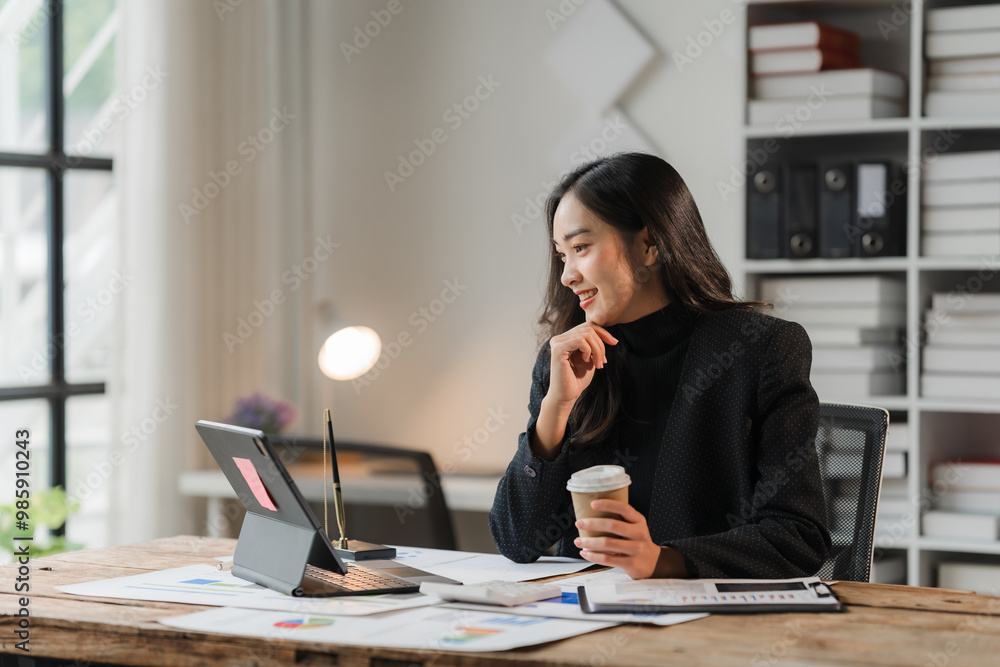 Confident Businesswoman: A young Asian businesswoman smiles confidently while reviewing documents and sipping coffee in a modern office setting. She embodies professionalism, focus, and success. Confident Businesswoman: A young Asian businesswoman smiles confidently while reviewing documents and sipping coffee in a modern office setting. She embodies professionalism, focus, and success.