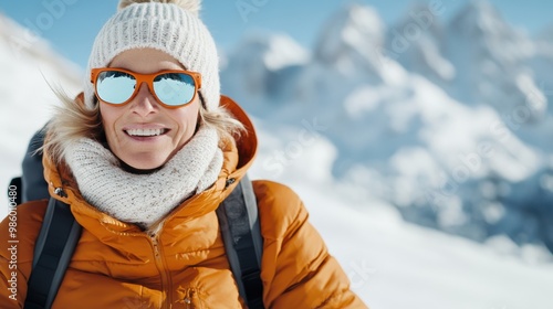 An individual dressed in a warm orange jacket, white knit hat, and fluffy scarf poses on a snowy mountain slope with a breathtaking alpine landscape in the background.