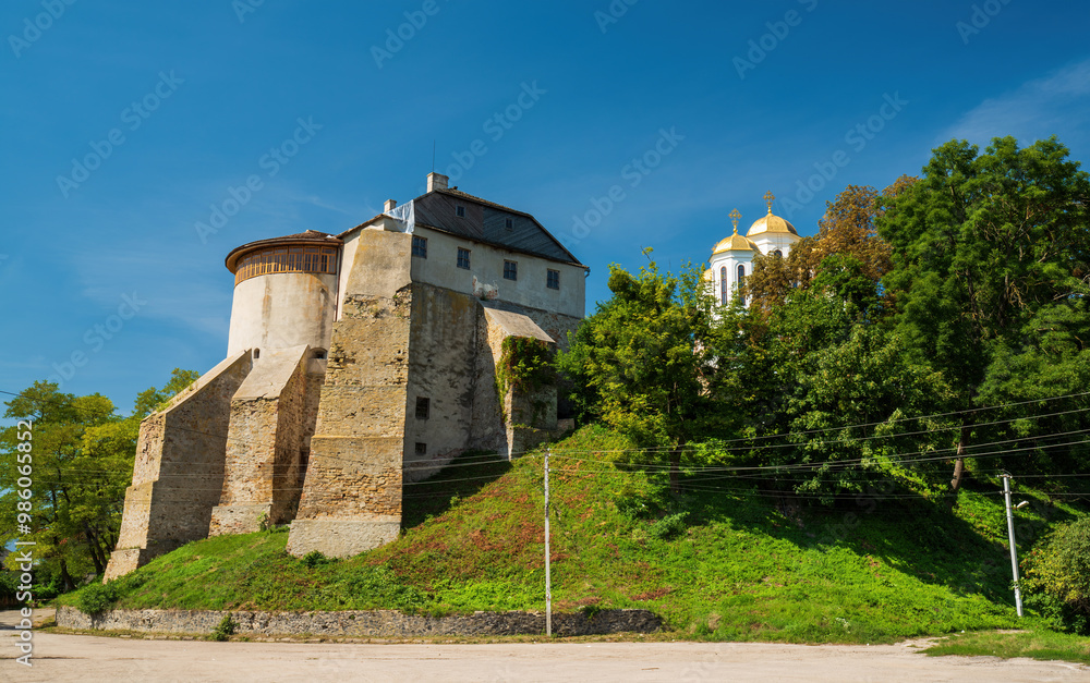 Fototapeta premium Ancient Ostroh Castle with its round tower stands on a green hill, alongside golden-domed Orthodox church. The historic fortress in Rivne region, Ukraine, basks in bright sunlight.