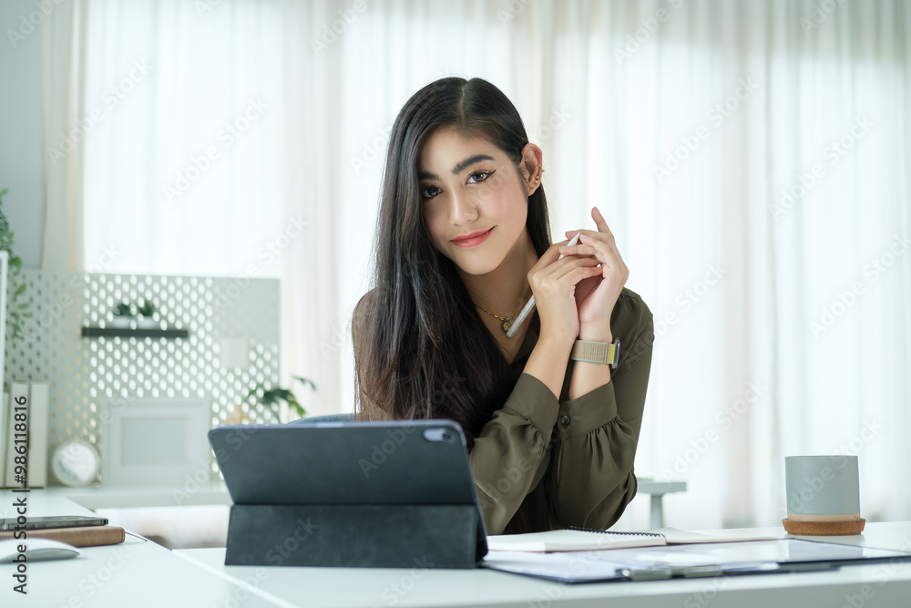 Confident young woman with long dark hair is seated at a sleek, modern desk, holding a stylus in her hand and smiling softly at the camera.