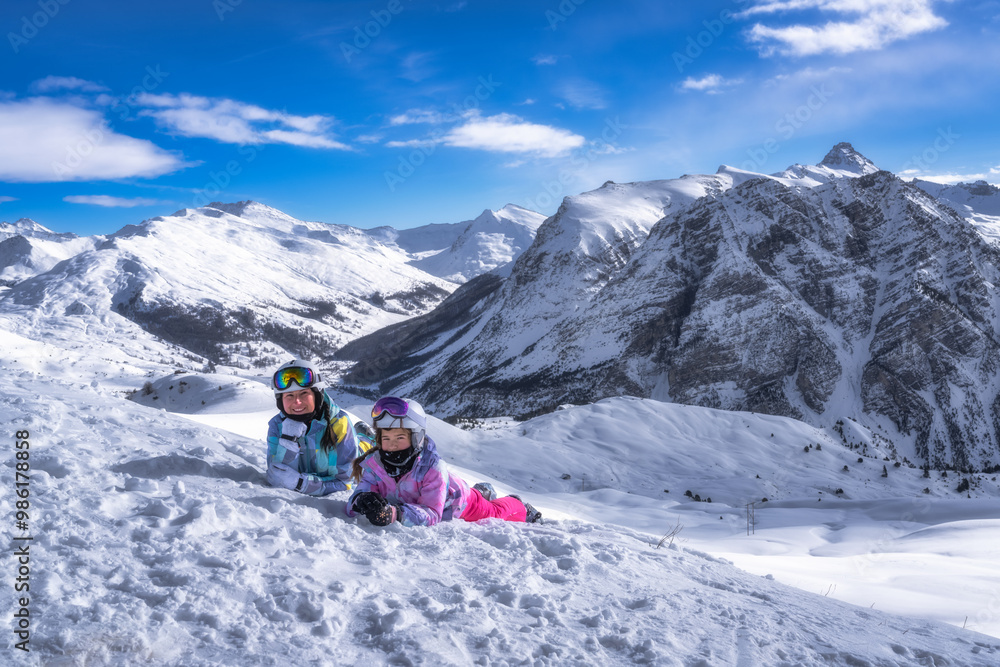 Mother and daughter in ski gear lying on snow, smiling and looking at camera, beautiful mountains in background, Montgenevre ski village, France, Alps Mother and daughter in ski gear lying on snow, smiling and looking at camera, beautiful mountains in background, Montgenevre ski village, France, Alps