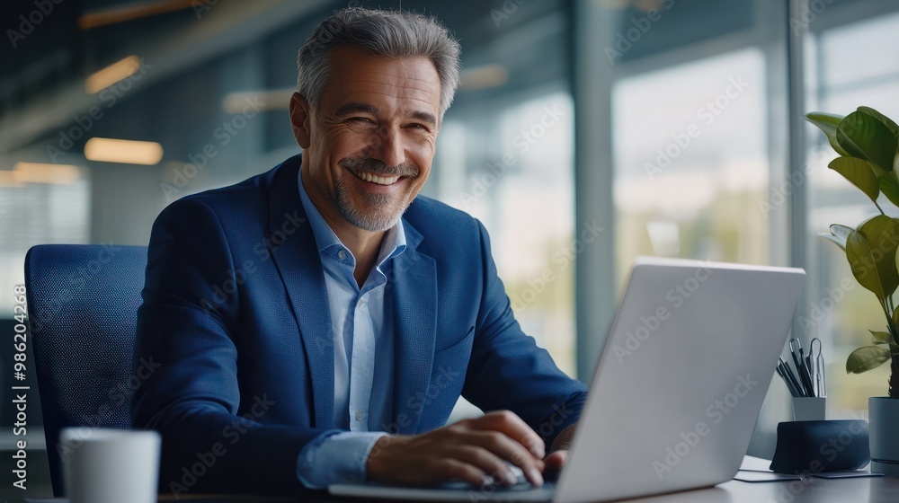 Happy smiling middle aged professional business man company executive ceo manager wearing blue suit sitting at desk in office working on laptop computer laughing at workplace. Portrait. Happy smiling middle aged professional business man company executive ceo manager wearing blue suit sitting at desk in office working on laptop computer laughing at workplace. Portrait.