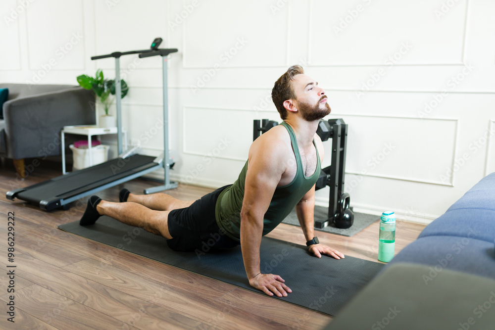 Fototapeta premium Athletic man doing yoga in cobra pose, stretching muscles on exercise mat in his home