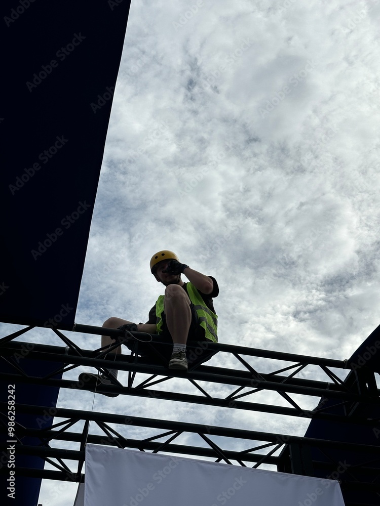 Young man in a green work vest and yellow helmet, perched on a metal construction framework. This photo highlights essential safety gear and the significance of safety in construction environments.