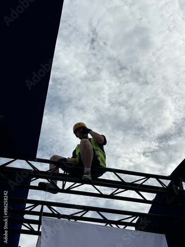 Obraz na płótnie Young man in a green work vest and yellow helmet, perched on a metal construction framework. This photo highlights essential safety gear and the significance of safety in construction environments. w salonie