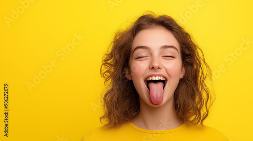 A young woman with brown curly hair sticks out her tongue and smiles at the camera against a bright yellow background.
