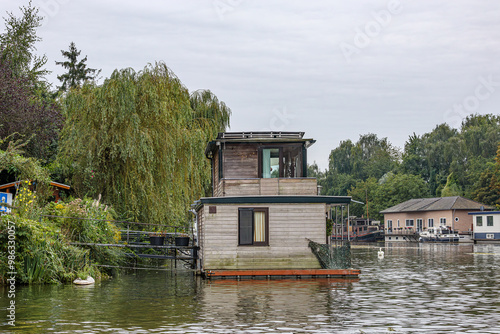 Houseboat moored in shipping canal outside city of Maastricht, wooden walls, small metal bridge, floating cityscape and leafy trees in background, cloudy day in South Limburg, Netherlands photo