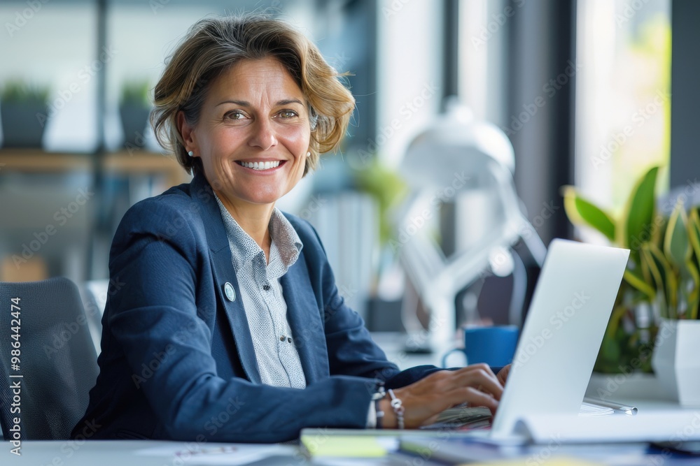 Happy smiling middle aged professional business woman company executive ceo manager wearing blue suit sitting at desk in office working on laptop computer Happy smiling middle aged professional business woman company executive ceo manager wearing blue suit sitting at desk in office working on laptop computer