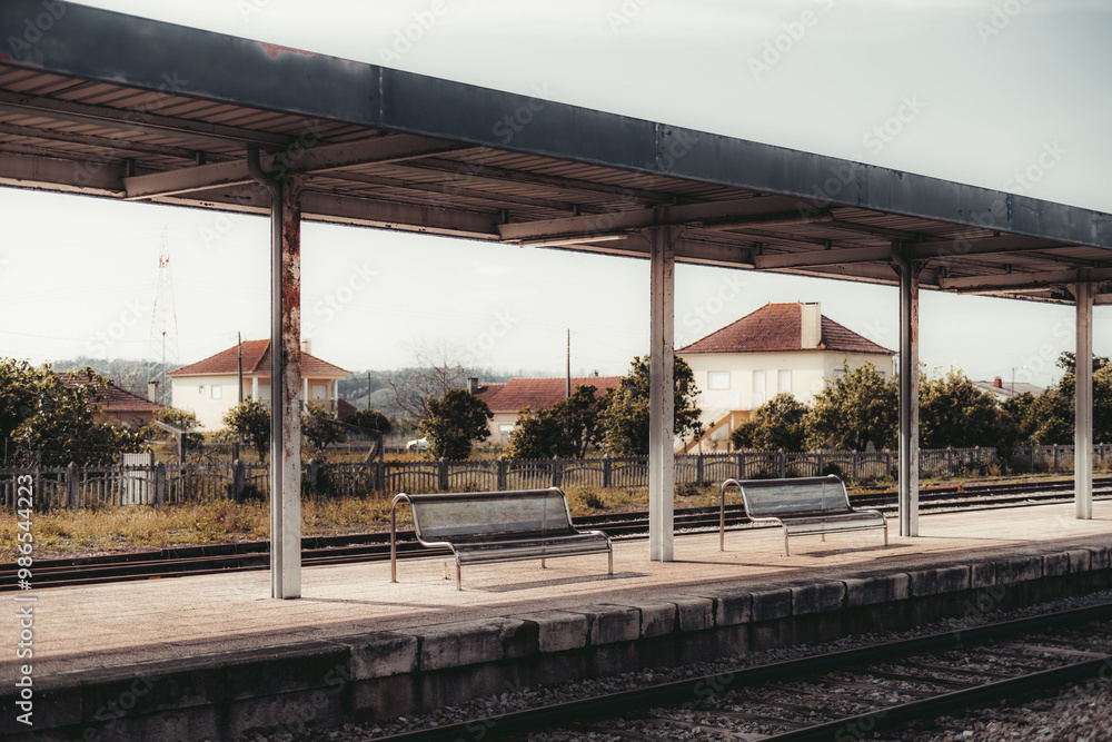 An empty train station platform with a metal roof and metal benches, located in a small rural area. Background shows houses with red tiled roofs and a wooden fence surrounding the property An empty train station platform with a metal roof and metal benches, located in a small rural area. Background shows houses with red tiled roofs and a wooden fence surrounding the property