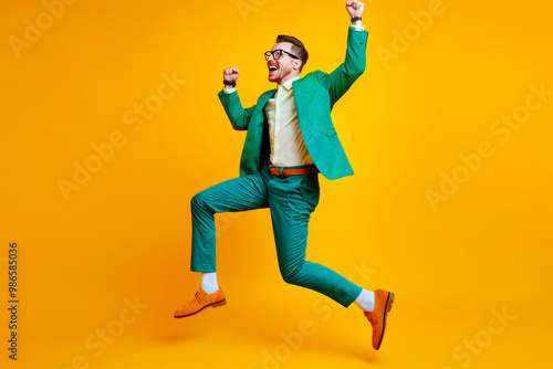 Young man celebrating success. Happy funny joyful excited guy in stylish green party suit and cool glasses raising fist up and dancing isolated on bright yellow background. Full length shot, side view