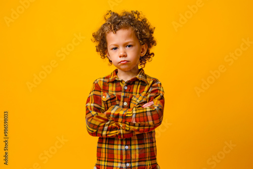 Full body serious kid model in trendy outfit keeping hands on waist and looking at camera against yellow background