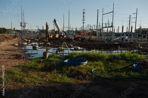 Wide view of the Electric substation under construction. Dirt piles from construction and equipment tire tracks with High Power Lines in Florida. Reflections in blue water. Blue sky.  Upgrading infras