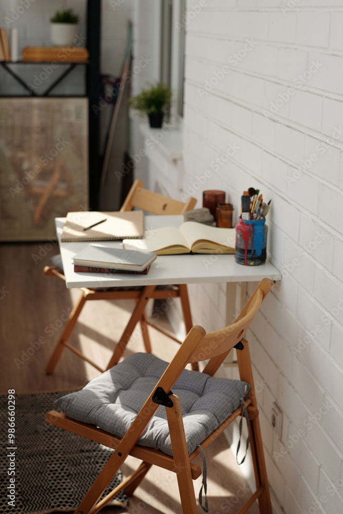 Sunlit office space featuring wooden chairs with cushions, white desk, and open notebooks. Potted plants and various writing tools adding a natural touch Sunlit office space featuring wooden chairs with cushions, white desk, and open notebooks. Potted plants and various writing tools adding a natural touch