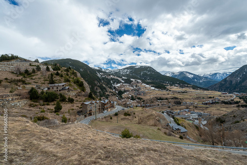 A mountain valley in Andorra reveals a picturesque town nestled among the slopes, with snow-capped peaks in the distance under a dramatic sky with swirling clouds. photo