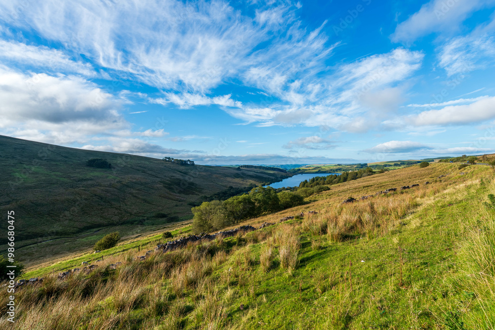 Naklejka premium Scenic Irish countryside with rolling hills, lake, and dramatic cloudscape on sunny day.