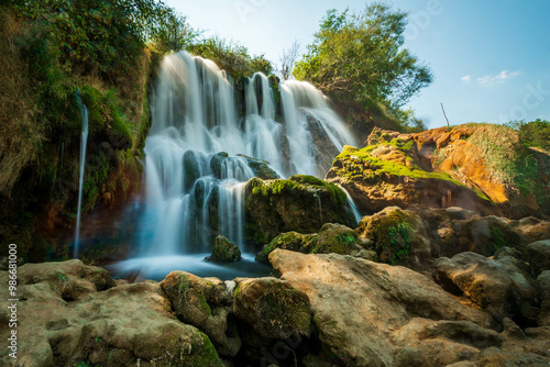 Kravice Waterfalls' cascading beauty is captured in a sunlit scene, showcasing its flowing waters and the vibrant textures of the surrounding rocks and moss. photo