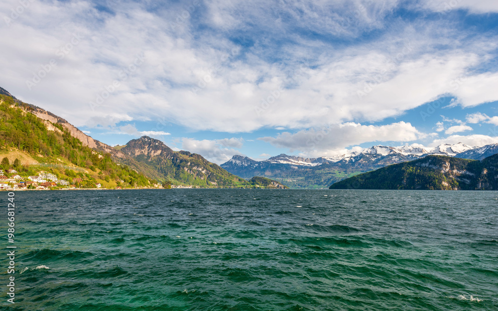 The deep green waters of Lake Lucerne in Switzerland are framed by verdant slopes and majestic snow-capped mountains under a dynamic sky with fluffy clouds, creating a quintessential Swiss landscape. The deep green waters of Lake Lucerne in Switzerland are framed by verdant slopes and majestic snow-capped mountains under a dynamic sky with fluffy clouds, creating a quintessential Swiss landscape.