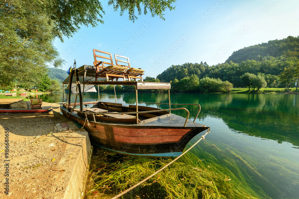 Obraz premium A traditional wooden boat rests peacefully on the serene Una River, surrounded by lush greenery and reflecting the clear blue sky, offering a tranquil escape in the heart of nature
