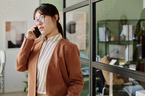 Woman standing in office, holding phone, wearing glasses and blazer, and having conversation. Modern office setup with glass partitions, shelves, and wall art