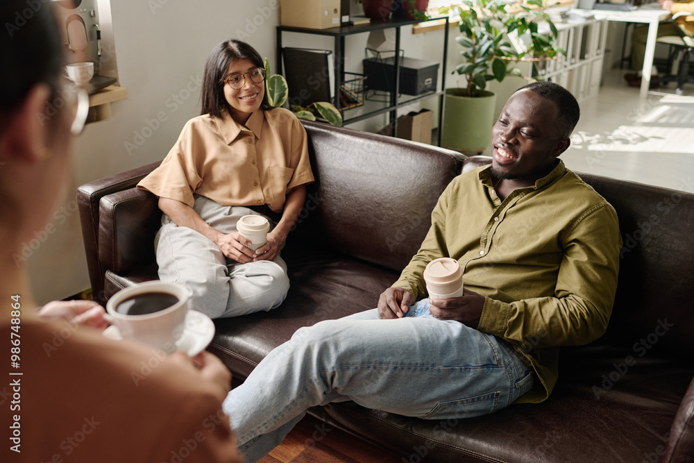 Obraz premium People sitting on comfortable couches enjoying coffee and casual conversation in a modern office environment with plants and natural light streaming in through large windows