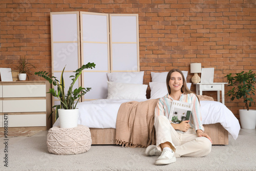 Young woman reading interior design magazine in cozy bedroom with brick wall ...