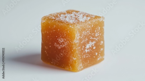 A single, square-shaped salted caramel candy cube rests on a pristine white background. Its golden hue and glistening surface suggest a rich, buttery flavor, while the scattering of salt crystals hint