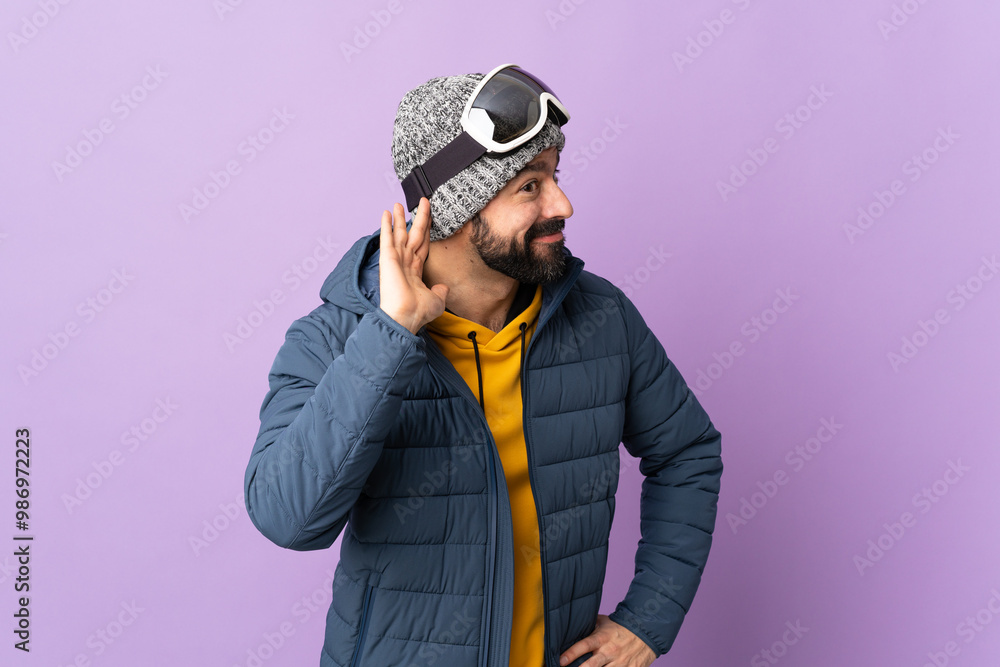 Skier man with snowboarding glasses over isolated purple background listening to something by putting hand on the ear Skier man with snowboarding glasses over isolated purple background listening to something by putting hand on the ear