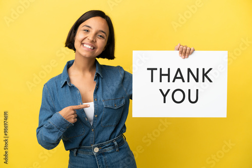 Young mixed race woman isolated on yellow background holding a placard with text THANK YOU and  pointing it