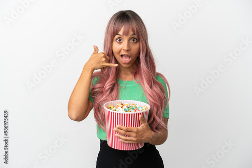 Young mixed race woman with pink hair eating popcorn isolated on white background making phone gesture. Call me back sign