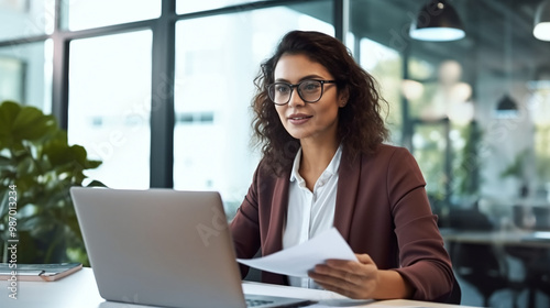 A female professional sits at her desk in an office, working on a laptop.