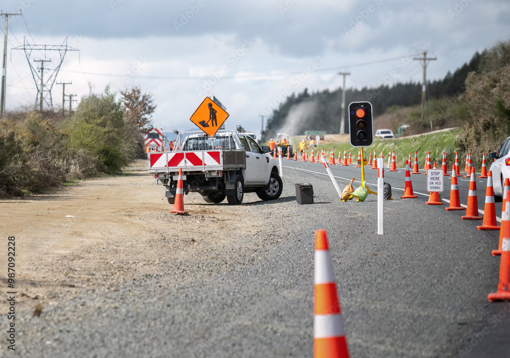 Orange traffic cones on the road. Cars stopped at the roadwork red light. Unrecognizable workers on the road.
