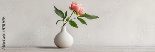 A modern botanical arrangement featuring a small white vase with a single stem of green foliage and a pink peony