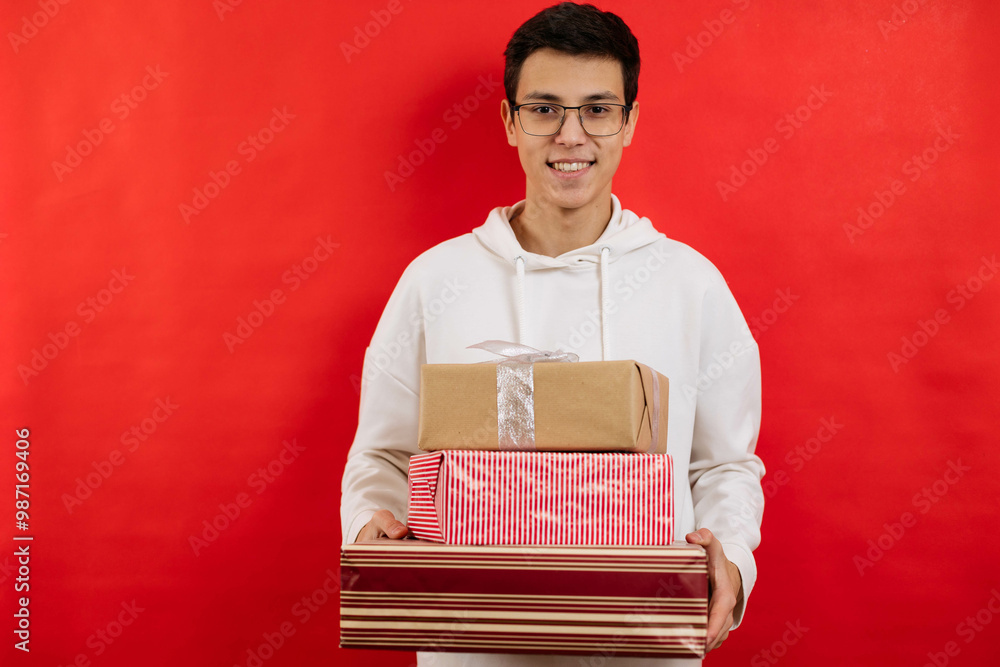 Cute young guy holding a box with a gift on a red background, smiling and looking at the camera. Cute young guy holding a box with a gift on a red background, smiling and looking at the camera.
