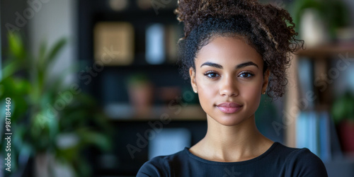 Young woman with curly hair standing confidently in a modern indoor space surrounded by plants and books in the afternoon light