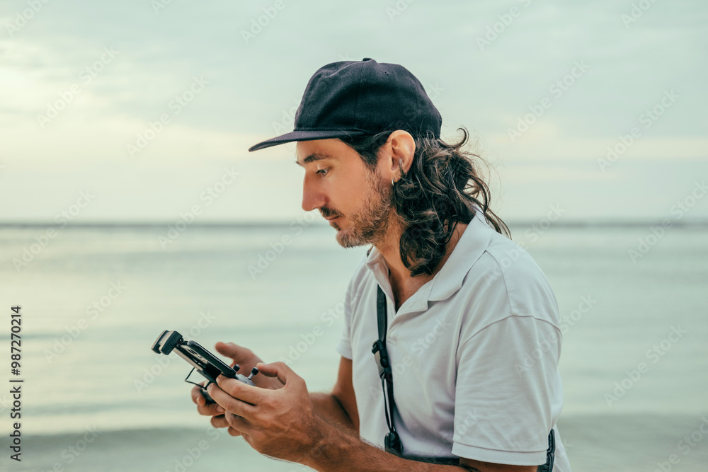 Obraz premium young handsome man with a beard and a cap controls a drone through a remote control, flight operator, FPV, control, video shooting, video make-up, concept, content, blogger, Thailand, sea