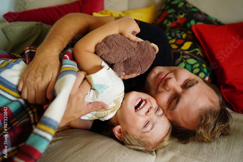 A joyful father and young son cuddling together on a bed, sharing a heartwarming moment with laughter and a stuffed toy.
 photo