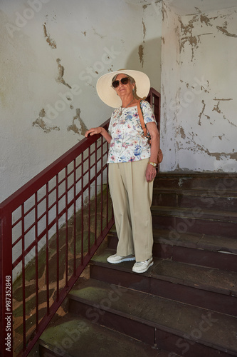 An octogenarian woman wearing sunglasses, a sun hat and a shoulder bag walks down a staircase with steel handrails and peeling walls. photo