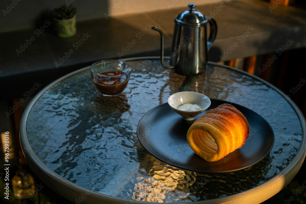 Kurtoskalacs, hungarian sweet chimney cake on a black plate. Served as a breakfast alongside black coffee, in a cafe environment Kurtoskalacs, hungarian sweet chimney cake on a black plate. Served as a breakfast alongside black coffee, in a cafe environment