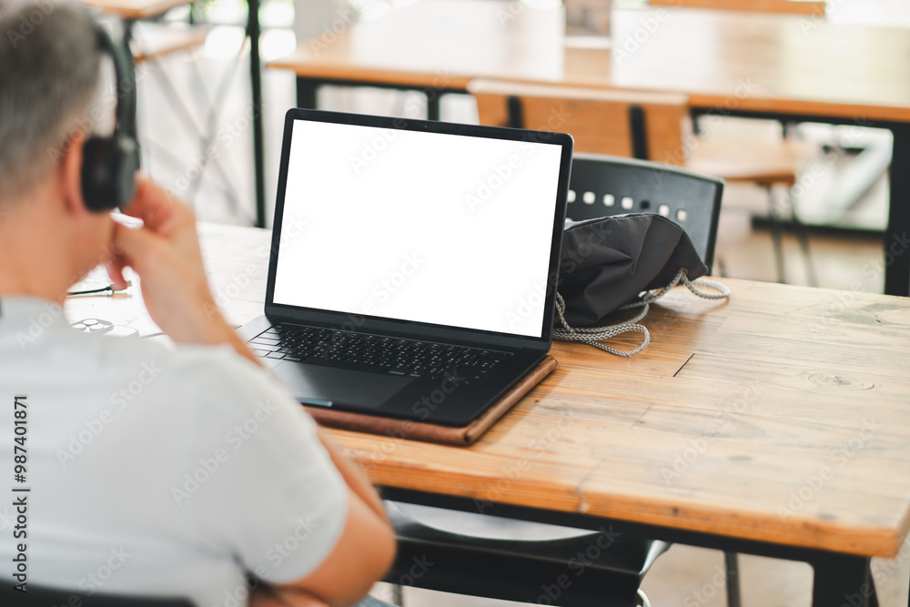 Man with headphones working on a laptop with a blank screen in a modern cafe. Wooden table and bright interior. Man with headphones working on a laptop with a blank screen in a modern cafe. Wooden table and bright interior.